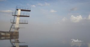 Verwitterter Sprungturm mit drei Ebenen, der sich im Nebel über einer stillen Wasserfläche abzeichnet.