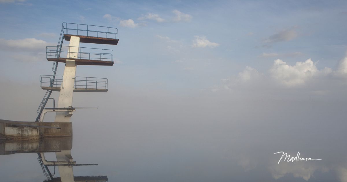Verwitterter Sprungturm mit drei Ebenen, der sich im Nebel über einer stillen Wasserfläche abzeichnet.