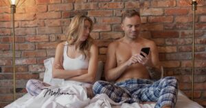 Frau und Mann sitzen nebeneinander im Bett vor einer Backsteinwand, sie mit verschränkten Armen zur Seite blickend, er auf sein Smartphone konzentriert.