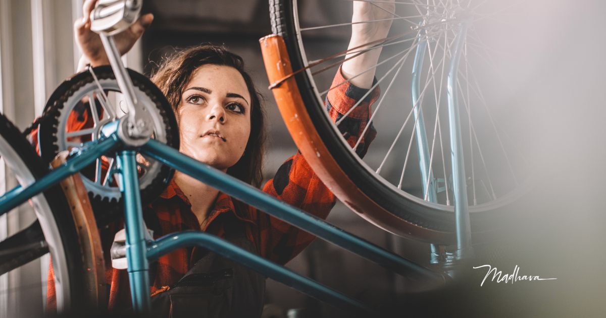 Eine Frau in kariertem Hemd arbeitet konzentriert an einem aufgebockten Fahrrad in einer Werkstatt, im Vordergrund Speichen und Kettenblatt.
